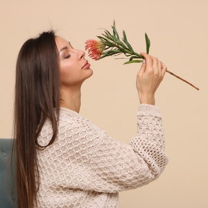 May include: A woman with long brown hair is wearing a cream-colored knitted sweater and smelling a red and green flower.