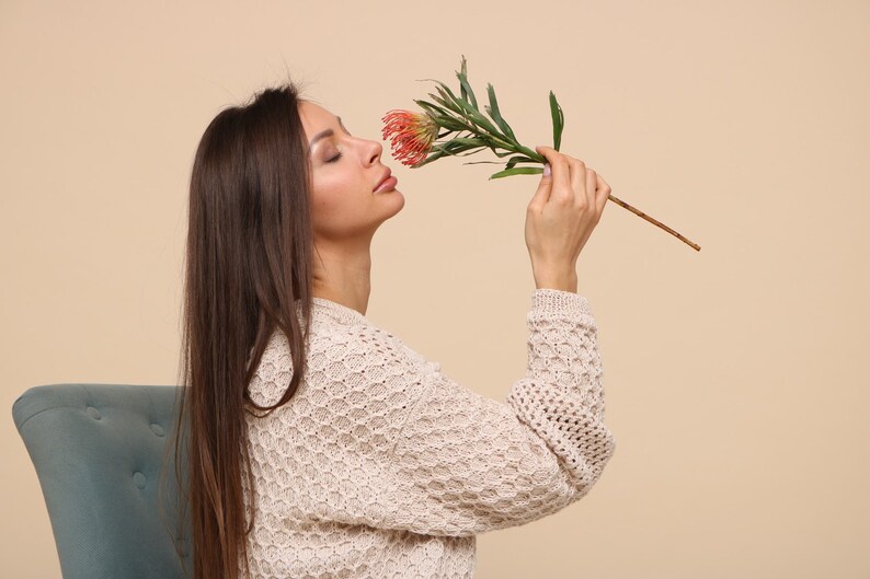 May include: A woman with long brown hair is wearing a cream-colored knitted sweater and smelling a red and green flower.