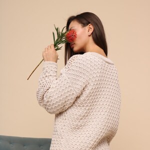 May include: A woman wearing a cream-colored, open-knit sweater with a honeycomb pattern. She is holding a red flower in front of her face.