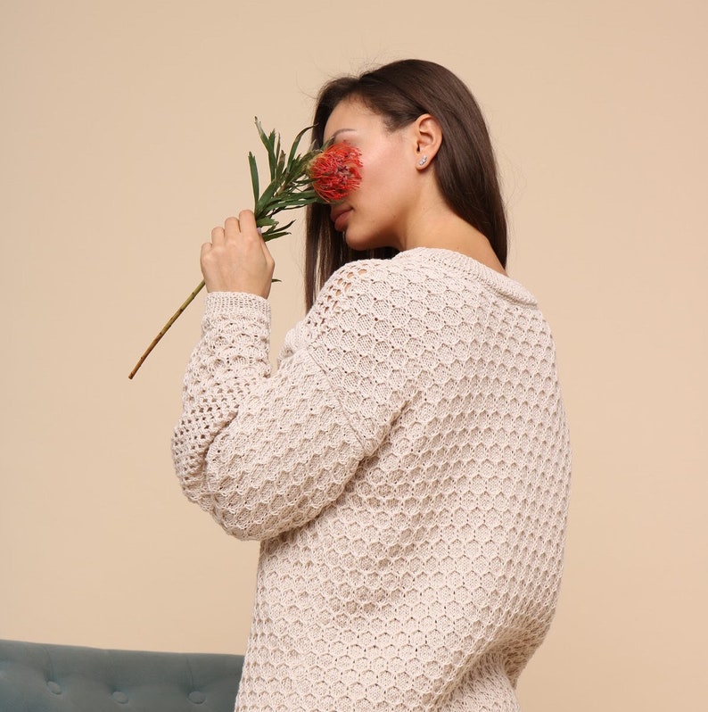 May include: A woman wearing a cream-colored, open-knit sweater with a honeycomb pattern. She is holding a red flower in front of her face.