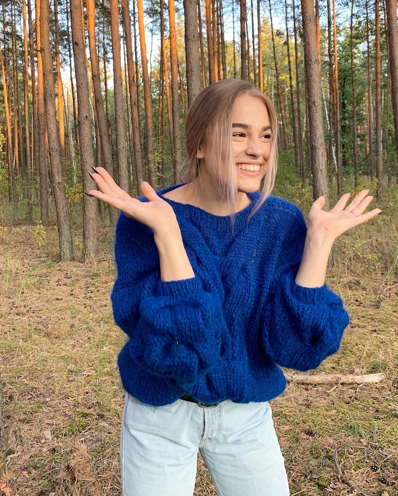 May include: A woman wearing a blue cable knit sweater and blue jeans smiles and raises her hands in the air. She is standing in a forest.