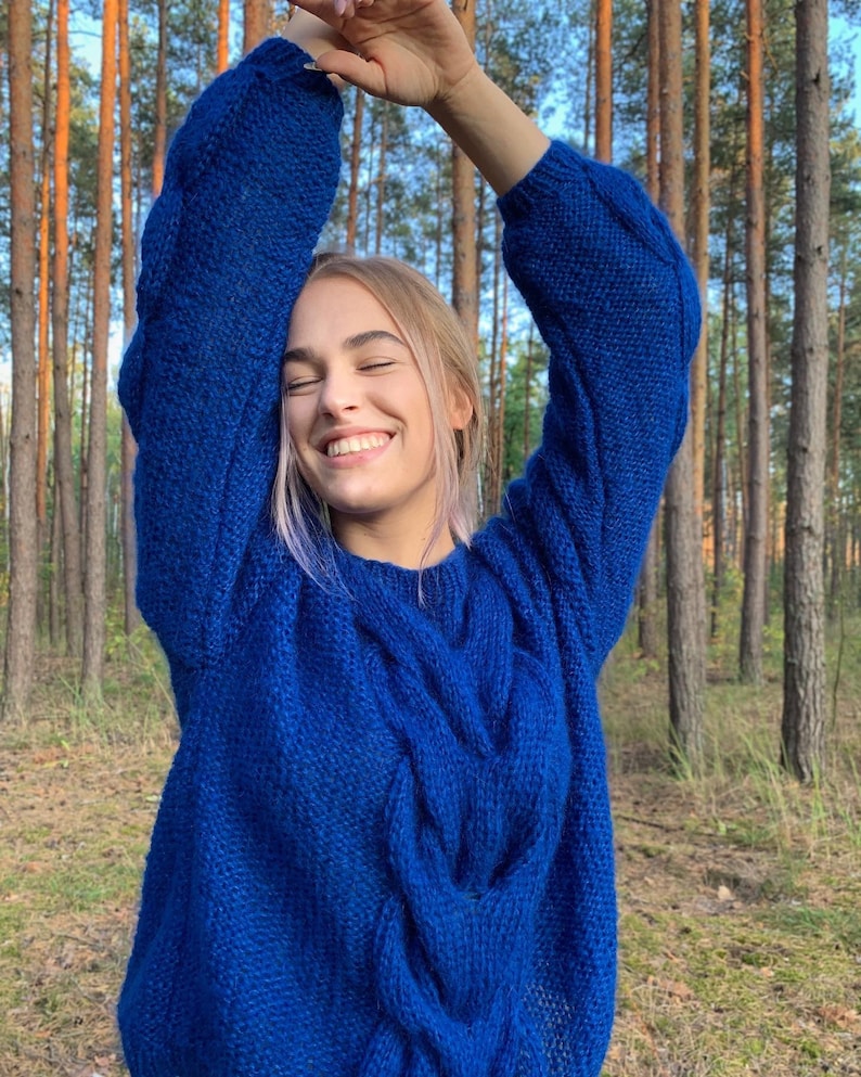 May include: A woman wearing a bright blue cable knit sweater with a relaxed fit. The sweater is made of a soft, fuzzy yarn.