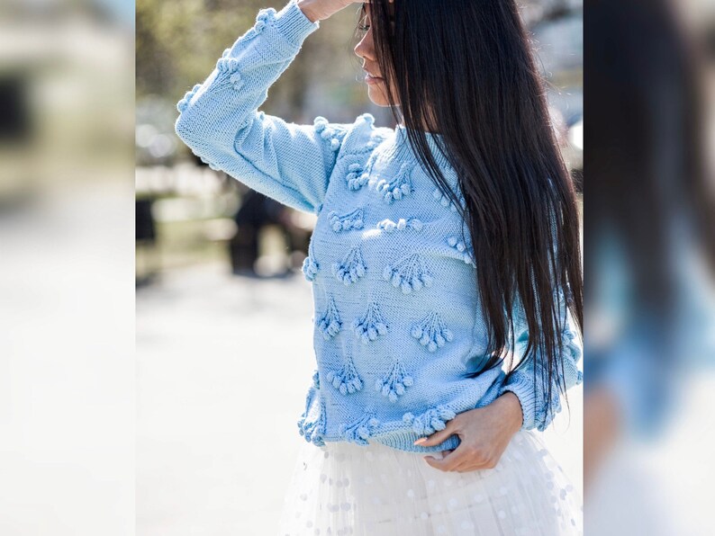May include: A light blue knitted sweater with a pom-pom design. The sweater has long sleeves and a round neckline.