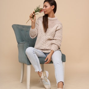 May include: A woman wearing light blue jeans and a cream-colored cable knit sweater sits on a blue velvet chair. She is holding a small sprig of pink flowers and smelling them.