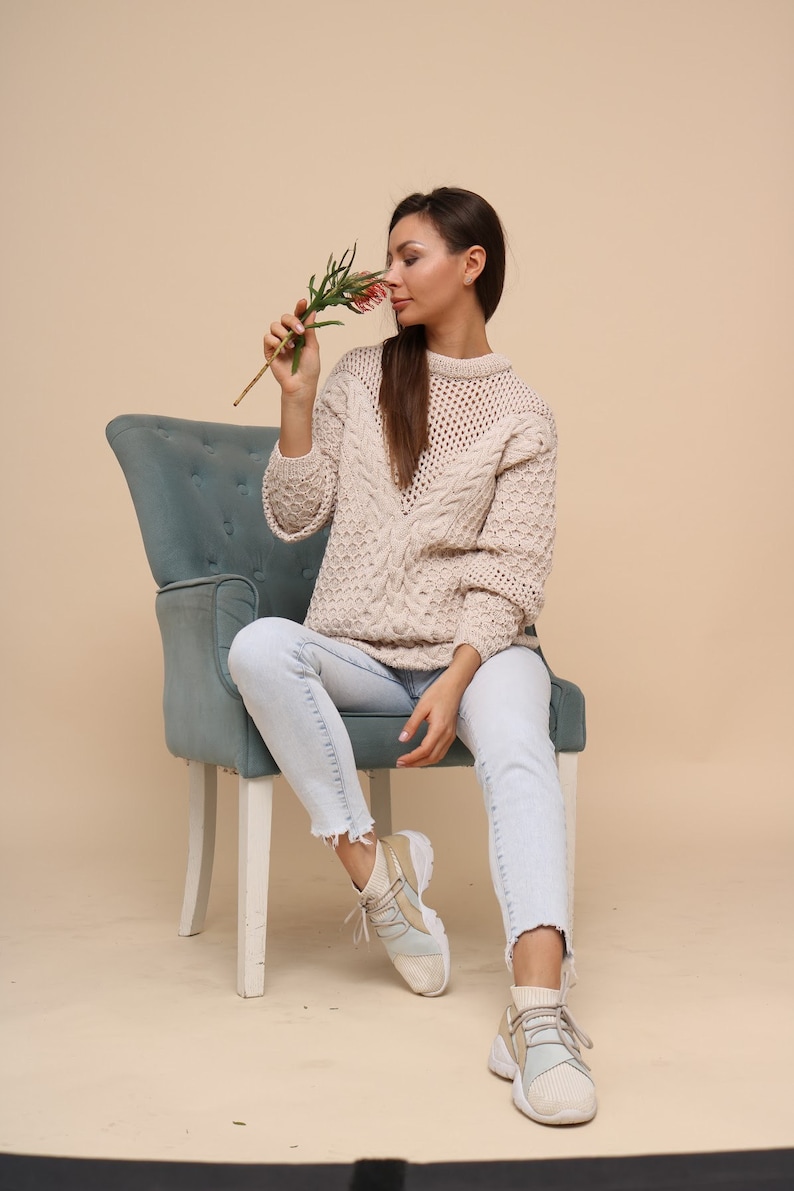 May include: A woman wearing light blue jeans and a cream-colored cable knit sweater sits on a blue velvet chair. She is holding a small sprig of pink flowers and smelling them.