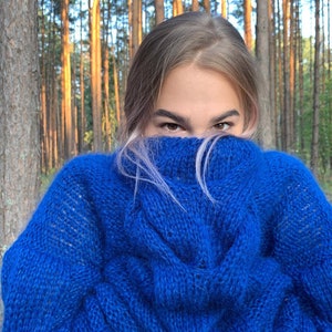 May include: A woman wearing a bright blue fuzzy sweater with a cable knit pattern. She is standing in a forest with tall trees behind her.