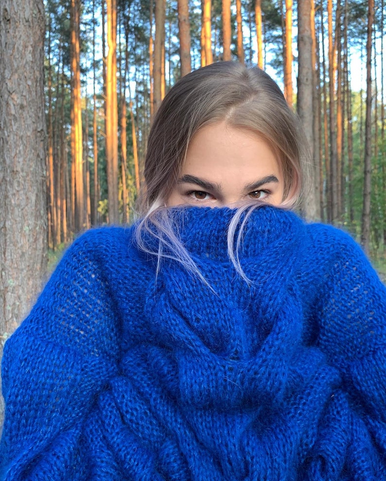 May include: A woman wearing a bright blue fuzzy sweater with a cable knit pattern. She is standing in a forest with tall trees behind her.