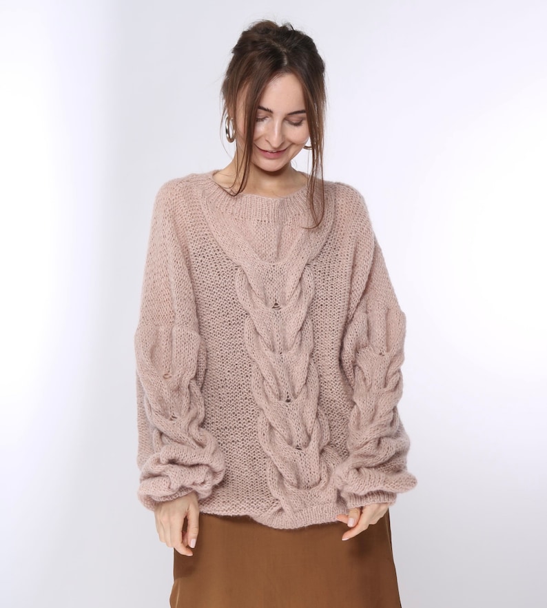 May include: A soft, blush-colored knitted sweater with a cable knit pattern and a relaxed fit.