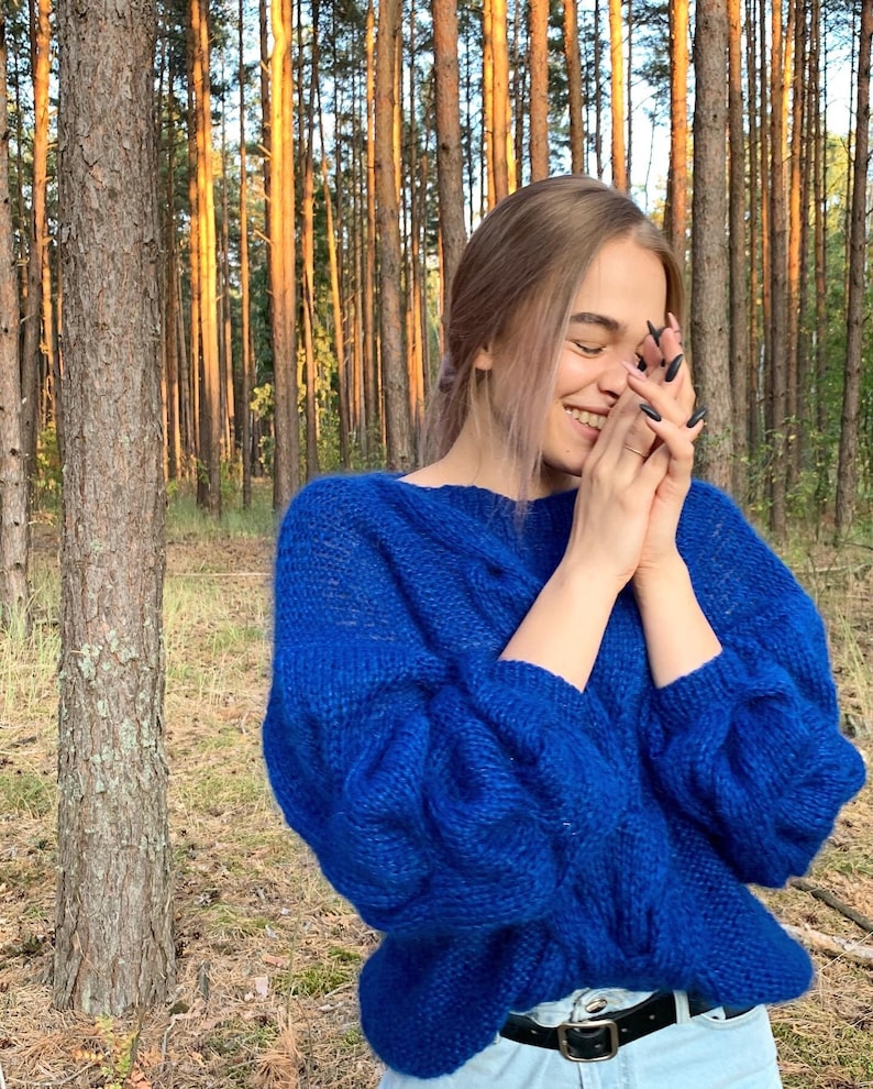 May include: A woman wearing a bright blue knitted sweater with long sleeves and a relaxed fit. She is standing in a forest with tall trees behind her.