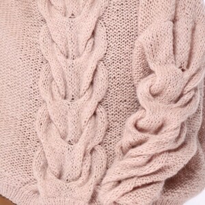 May include: A close-up of a soft, pale pink knitted sweater with a cable knit pattern. The sweater has long sleeves and a relaxed fit.