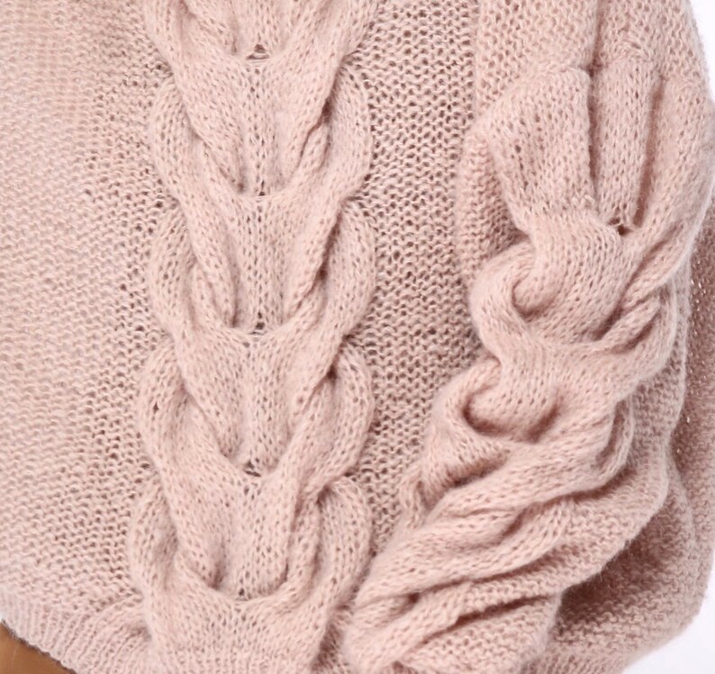 May include: A close-up of a soft, pale pink knitted sweater with a cable knit pattern. The sweater has long sleeves and a relaxed fit.