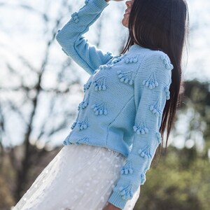 May include: A light blue sweater with a textured design and white pom-poms. The sweater is worn over a white tulle skirt with white polka dots.
