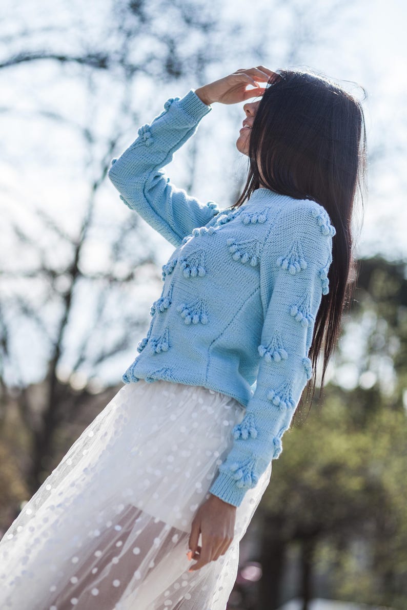 May include: A light blue sweater with a textured design and white pom-poms. The sweater is worn over a white tulle skirt with white polka dots.