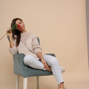 May include: A woman wearing a light beige knitted sweater and light blue jeans sits in a teal armchair with white legs. She is holding a red flower in front of her face.