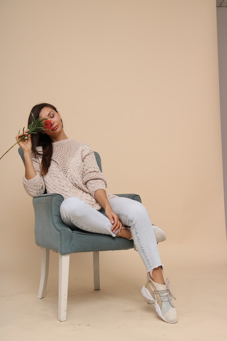 May include: A woman wearing a light beige knitted sweater and light blue jeans sits in a teal armchair with white legs. She is holding a red flower in front of her face.