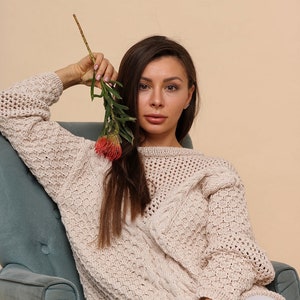 May include: A woman wearing a cream-colored knitted sweater with a cable knit design sits in a teal armchair. She holds a sprig of greenery with a red flower in her hand.