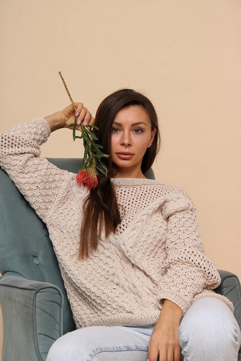 May include: A woman wearing a cream-colored knitted sweater with a cable knit design sits in a teal armchair. She holds a sprig of greenery with a red flower in her hand.