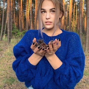 May include: A woman wearing a blue knitted sweater with a cable knit pattern. She is holding a handful of pine cones in her hands.