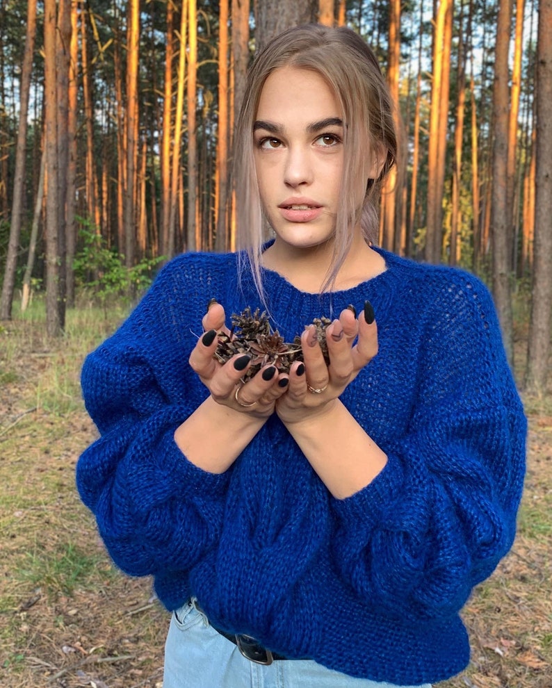 May include: A woman wearing a blue knitted sweater with a cable knit pattern. She is holding a handful of pine cones in her hands.
