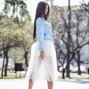 May include: A woman wearing a light blue sweater with white pom-poms and a white tulle skirt with polka dots. She is wearing nude heels and walking on a paved surface.