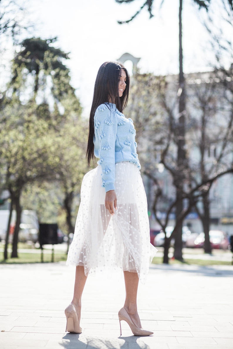 May include: A woman wearing a light blue sweater with white pom-poms and a white tulle skirt with polka dots. She is wearing nude heels and walking on a paved surface.