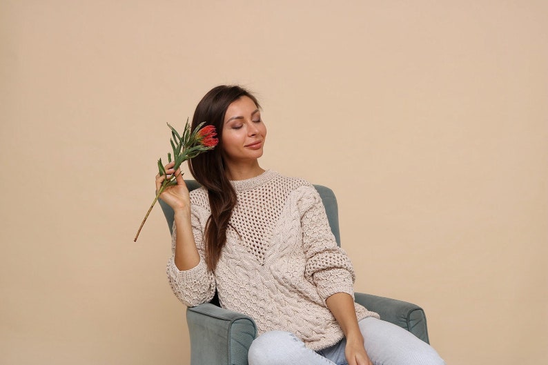 May include: A woman wearing a cream-colored knitted sweater sits in a teal armchair and holds a red flower with green leaves.
