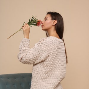 May include: A woman wearing a beige knitted sweater with a honeycomb pattern and blue jeans is smelling a pink flower.