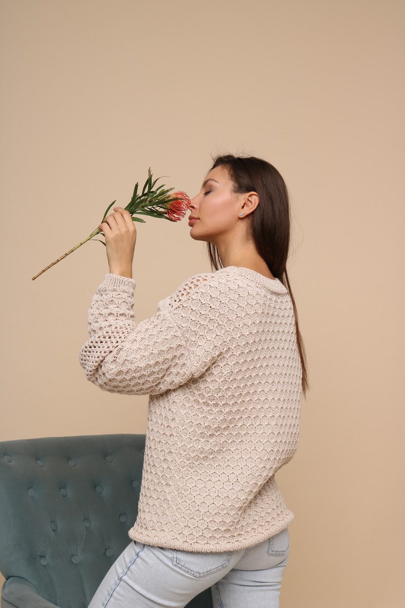 May include: A woman wearing a beige knitted sweater with a honeycomb pattern and blue jeans is smelling a pink flower.