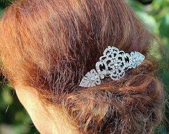 May include: A silver hair comb with a delicate, intricate design featuring clear rhinestones. The comb is worn in a woman's red hair.