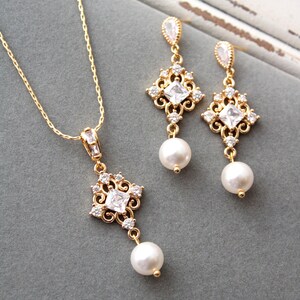 May include: A gold necklace and earrings set with a delicate, ornate design featuring a square-shaped crystal and a pearl drop. The jewelry is on a gray background.