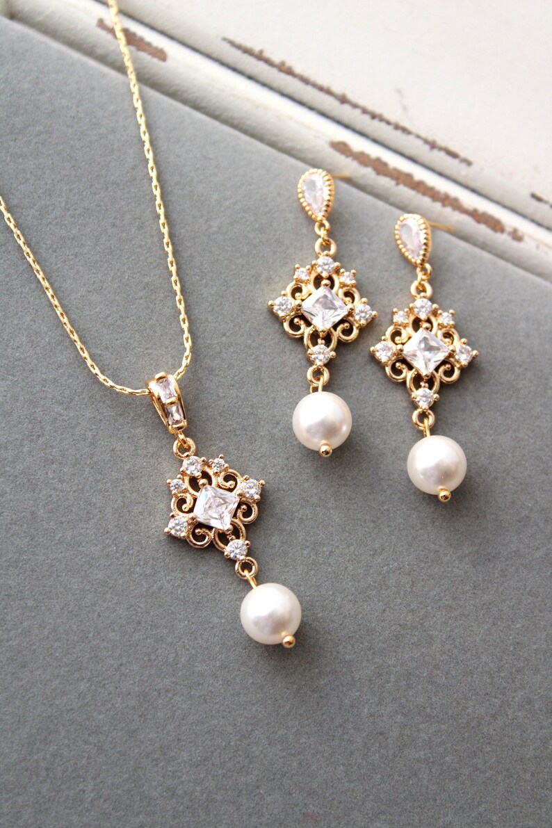May include: A gold necklace and earrings set with a delicate, ornate design featuring a square-shaped crystal and a pearl drop. The jewelry is on a gray background.
