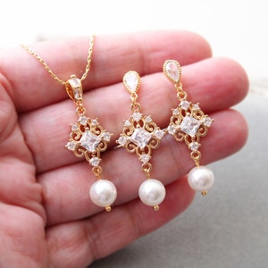 May include: A gold necklace and earrings set with a square-shaped crystal pendant and pearl drop. The pendant features a delicate filigree design.