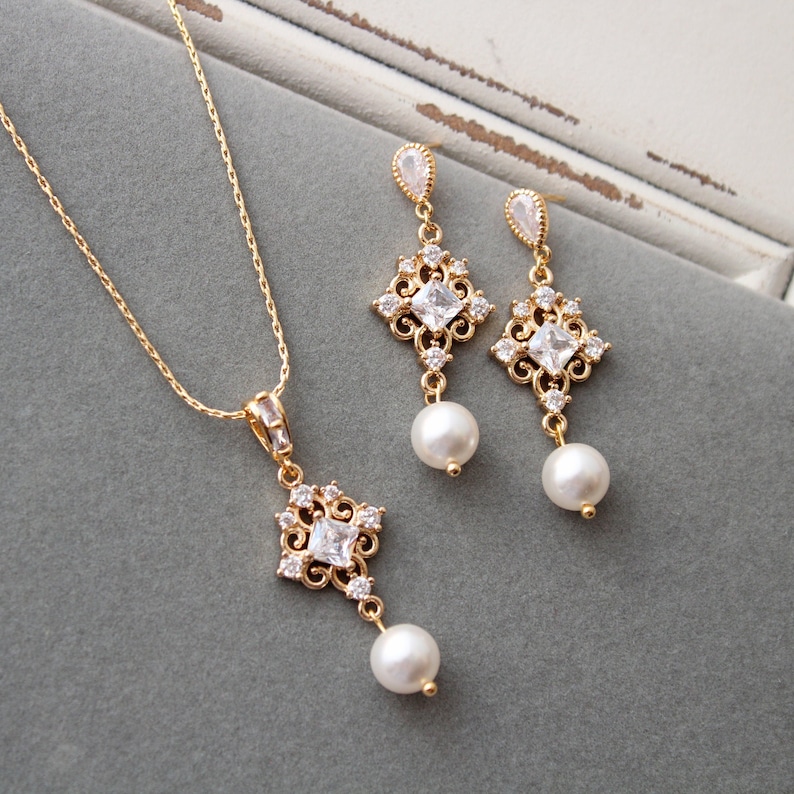May include: A gold necklace and earrings set with a square pendant and pearl drop. The pendant features a delicate filigree design with clear crystals.