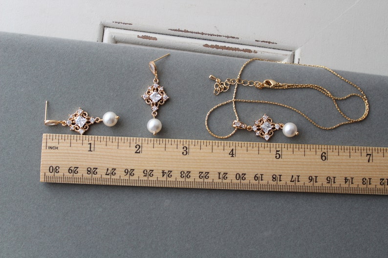 May include: A gold necklace and earrings set with a square pendant and pearl drop. The pendant is adorned with clear cubic zirconia stones. The necklace is 18 inches long and the earrings are 2 inches long.