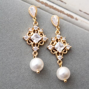 May include: Gold dangle earrings with a square-shaped crystal and a pearl drop. The earrings have a delicate, ornate design.