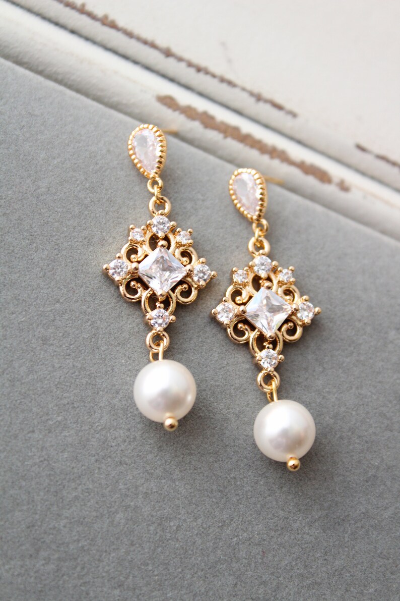 May include: Gold dangle earrings with a square-shaped crystal and a pearl drop. The earrings have a delicate, ornate design.