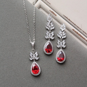 May include: A silver necklace and earrings set with a red teardrop-shaped gemstone surrounded by clear rhinestones. The necklace features a pendant with a cluster of clear rhinestones in the shape of leaves. The earrings are dangle earrings with a similar leaf design.