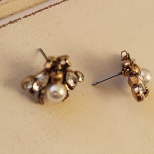 May include: A pair of gold bee stud earrings with a pearl accent. The earrings are made of metal and have a detailed design.