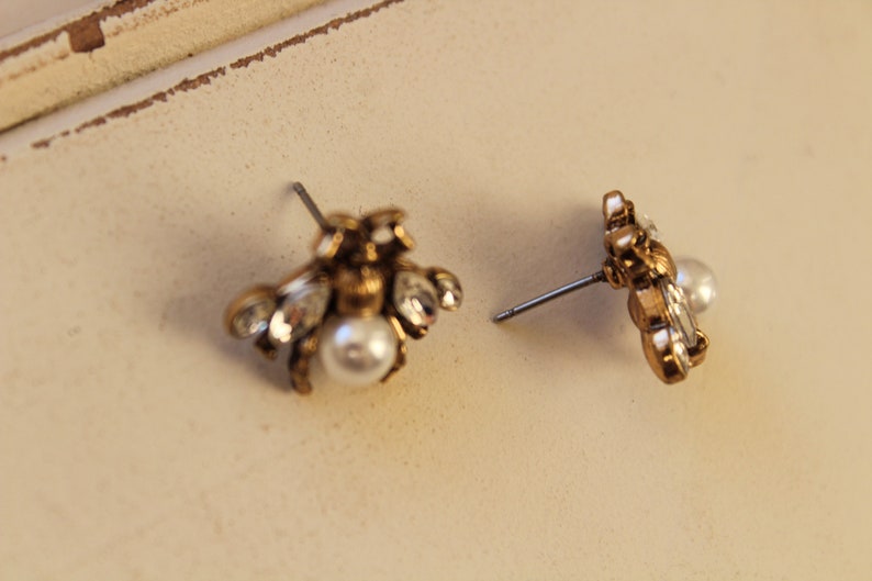 May include: A pair of gold bee stud earrings with a pearl accent. The earrings are made of metal and have a detailed design.