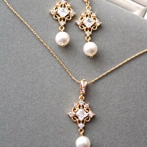 May include: A gold necklace and earrings set with a delicate chain and a square pendant with clear stones and a pearl drop. The earrings have a similar design with a pearl drop.