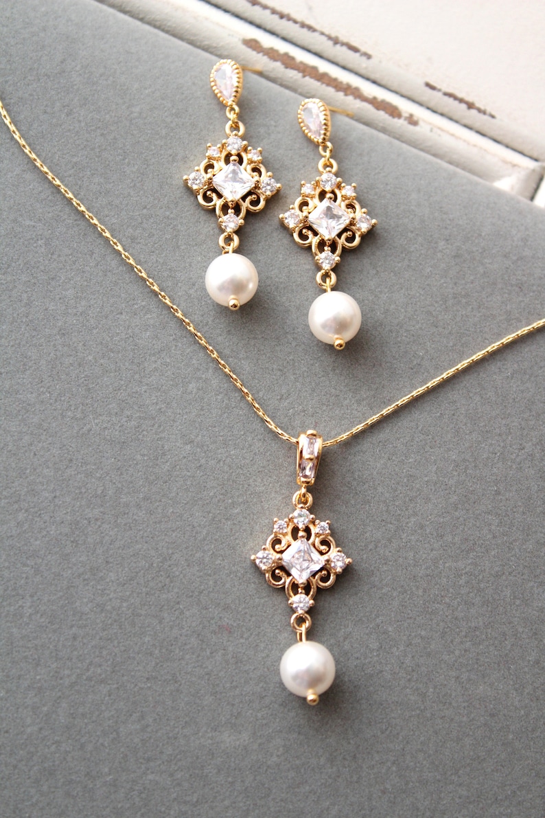 May include: A gold necklace and earrings set with a delicate chain and a square pendant with clear stones and a pearl drop. The earrings have a similar design with a pearl drop.
