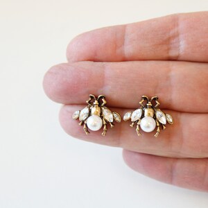 May include: A pair of gold bee stud earrings with a pearl center and clear crystal accents.