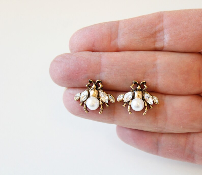 May include: A pair of gold bee stud earrings with a pearl center and clear crystal accents.