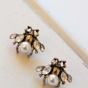 May include: A pair of gold-tone bee stud earrings with clear crystal accents and a white pearl center.