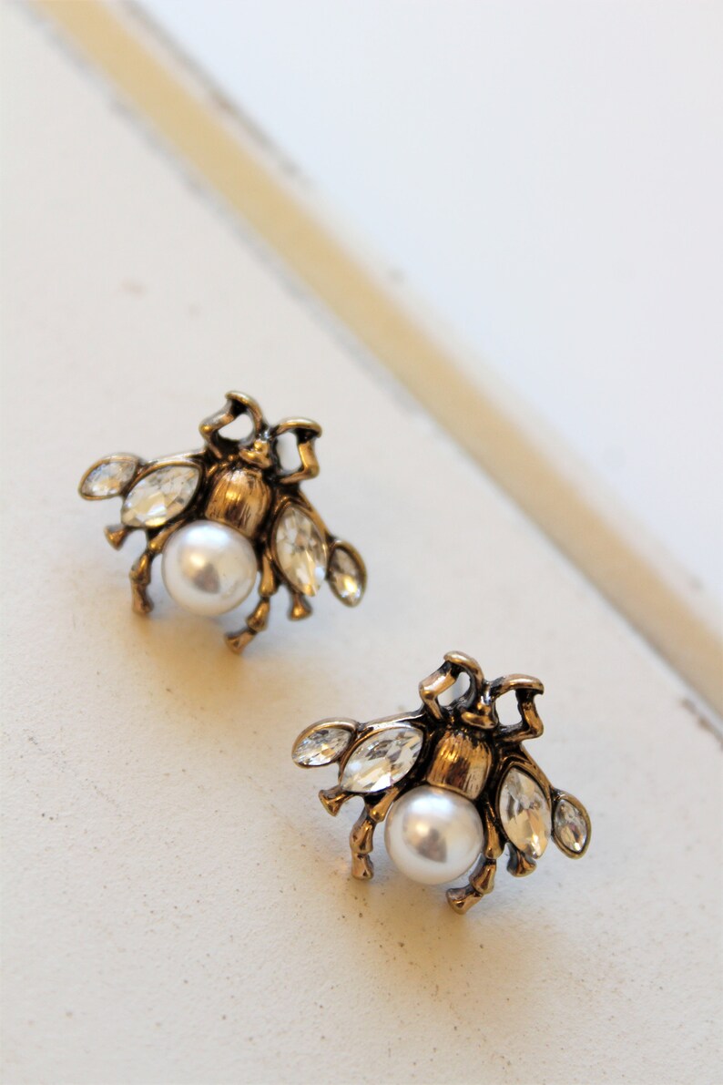 May include: A pair of gold-tone bee stud earrings with clear crystal accents and a white pearl center.
