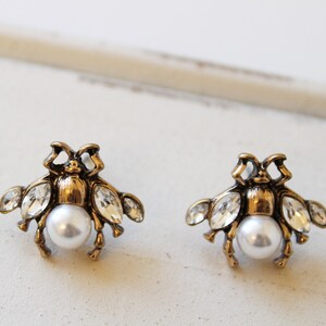 May include: A pair of gold-tone earrings featuring a bee design with clear crystal accents and a pearl center.