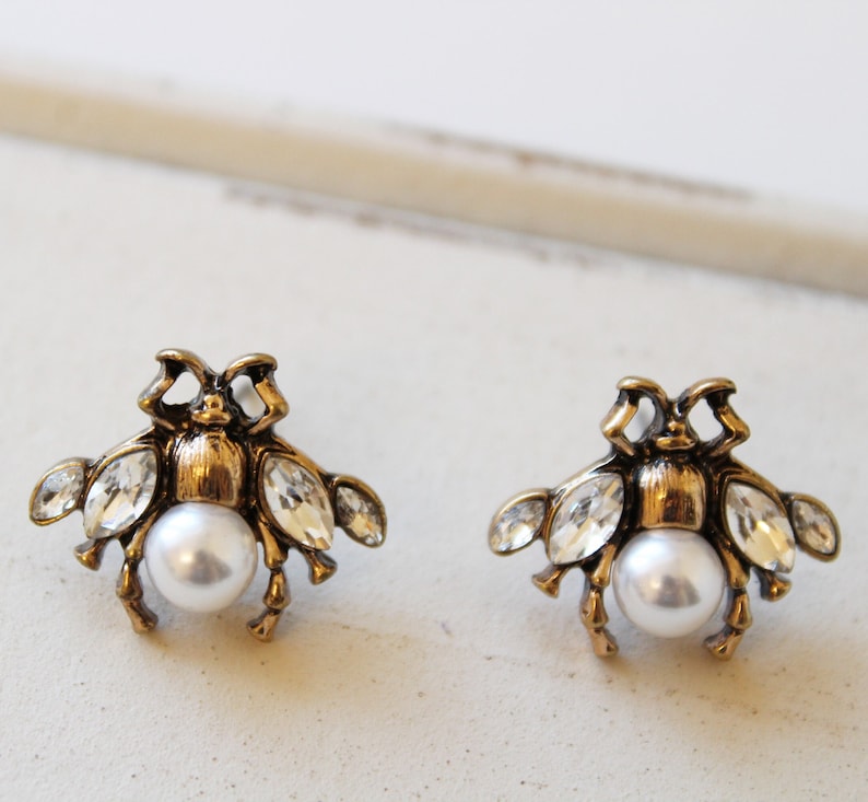 May include: A pair of gold-tone earrings featuring a bee design with clear crystal accents and a pearl center.