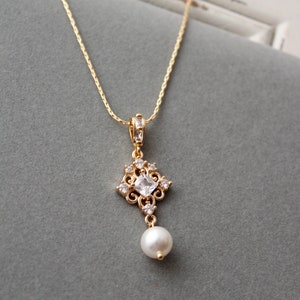 May include: A gold chain necklace with a pendant featuring a square-shaped clear crystal surrounded by smaller crystals and a single pearl drop.