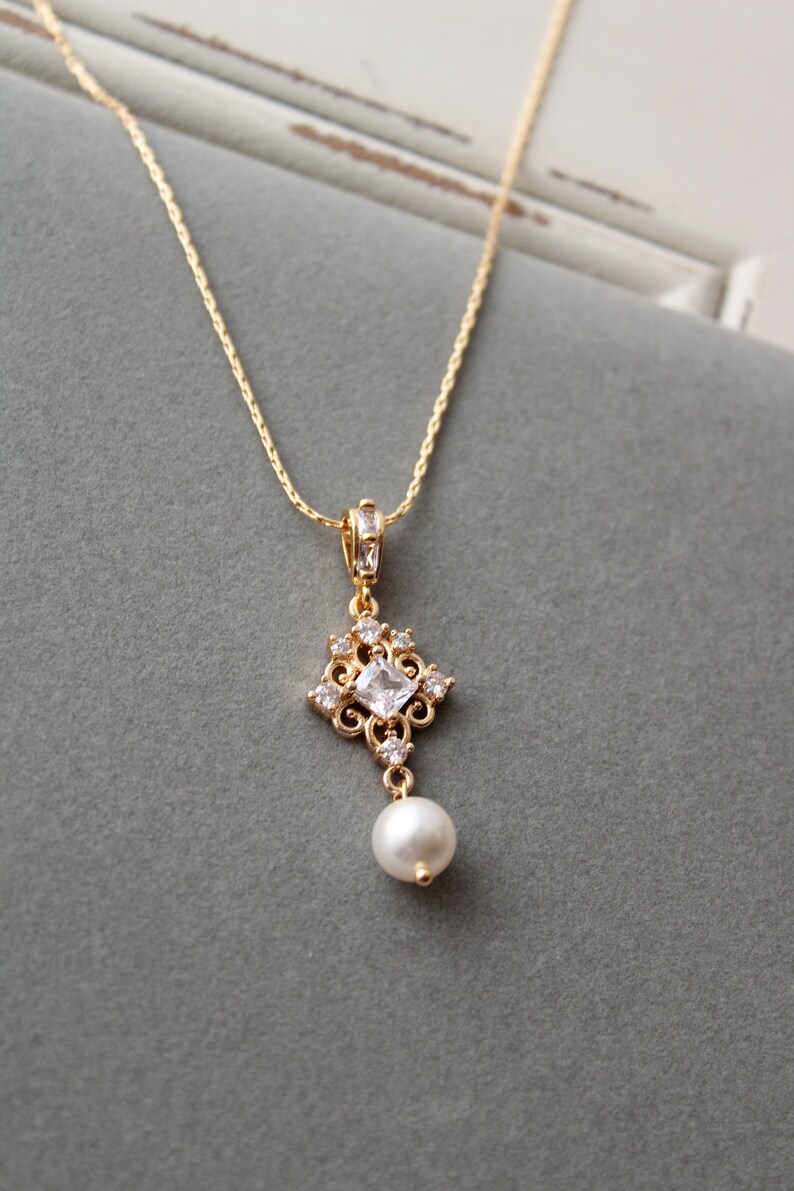 May include: A gold chain necklace with a pendant featuring a square-shaped clear crystal surrounded by smaller crystals and a single pearl drop.