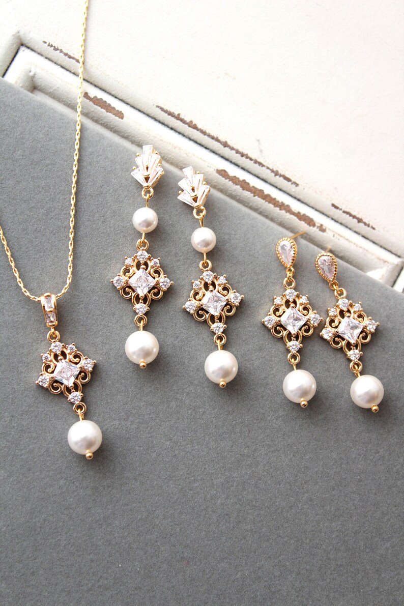 May include: A gold necklace and earrings set with a delicate chain, a square pendant with clear stones, and a pearl drop. The earrings have a similar design with a square pendant and a pearl drop.
